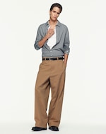 Placeholder for: Person in a checkered shirt and brown wide-leg pants stands against a plain background, holding their collar with one hand.