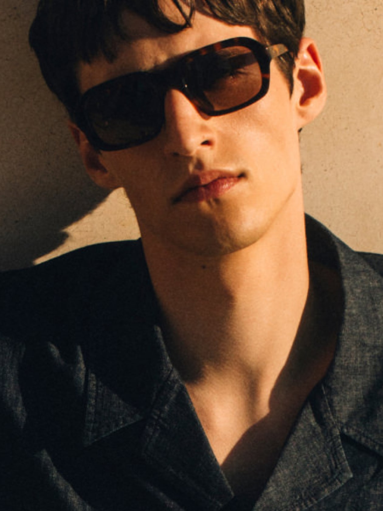 Person in sunglasses wearing a dark shirt, with short hair, leans against a sunlit wall, casting a shadow on the face.