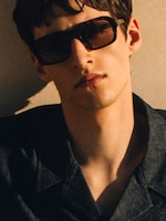 Placeholder for: Person in sunglasses wearing a dark shirt, with short hair, leans against a sunlit wall, casting a shadow on the face.
