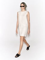 Placeholder for: A person wearing a sleeveless beige dress, black sunglasses, and a patterned headscarf, walking confidently in black sandals.