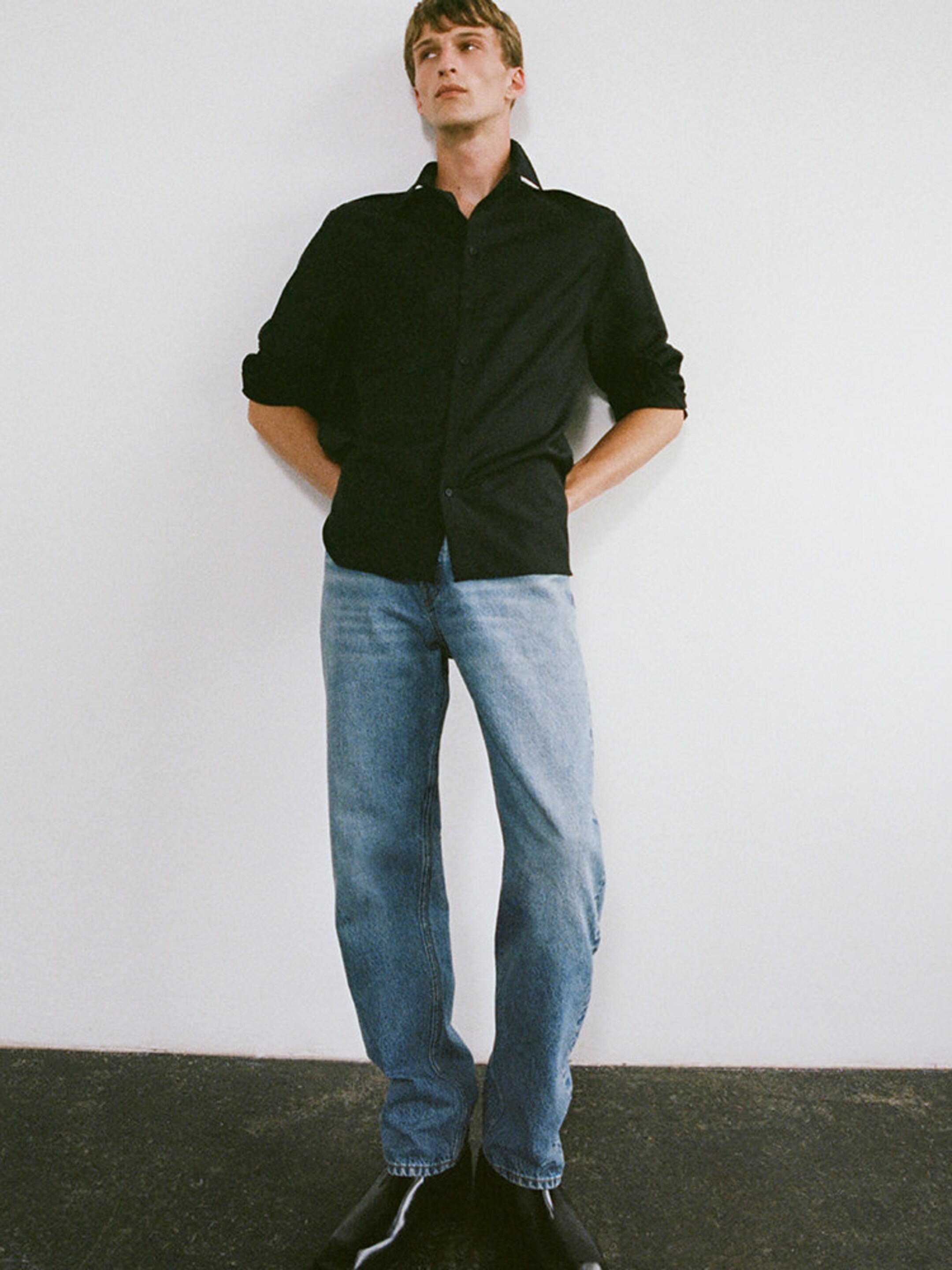 Person in a black shirt and blue jeans stands against a white wall with hands in pockets, wearing black boots.