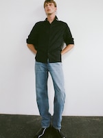 Placeholder for: Person in a black shirt and blue jeans stands against a white wall with hands in pockets, wearing black boots.