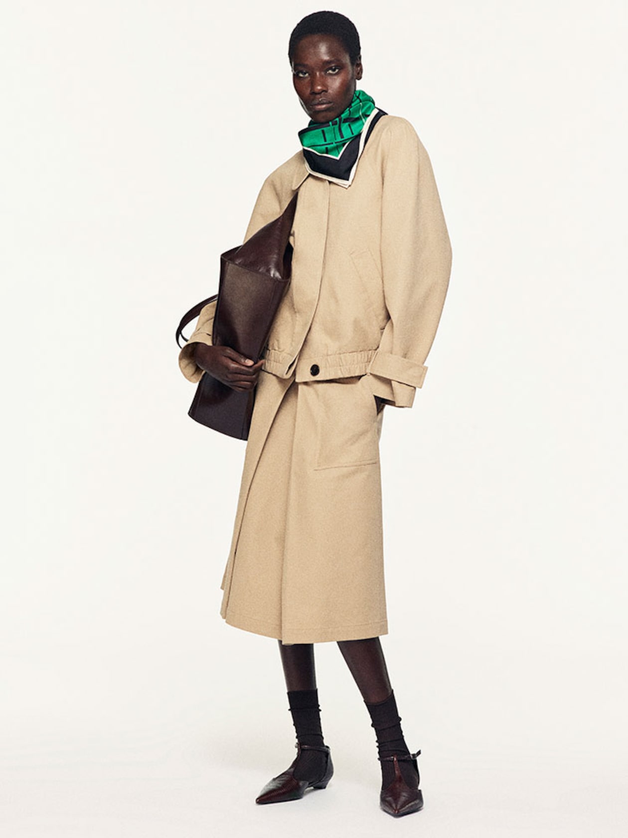A person wearing a tan outfit with a green scarf, holding a large brown bag, and dressed in dark ankle socks and pointed shoes.