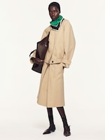 Placeholder for: A person wearing a tan outfit with a green scarf, holding a large brown bag, and dressed in dark ankle socks and pointed shoes.