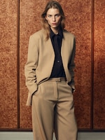 Placeholder for: Person wearing a beige suit and dark shirt stands against a textured brown background, hands in pockets, looking directly at the camera.