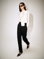 Placeholder for: Woman wearing a polka dot blouse, black high-waisted trousers, dark sunglasses, and pointed brown shoes, walking confidently.