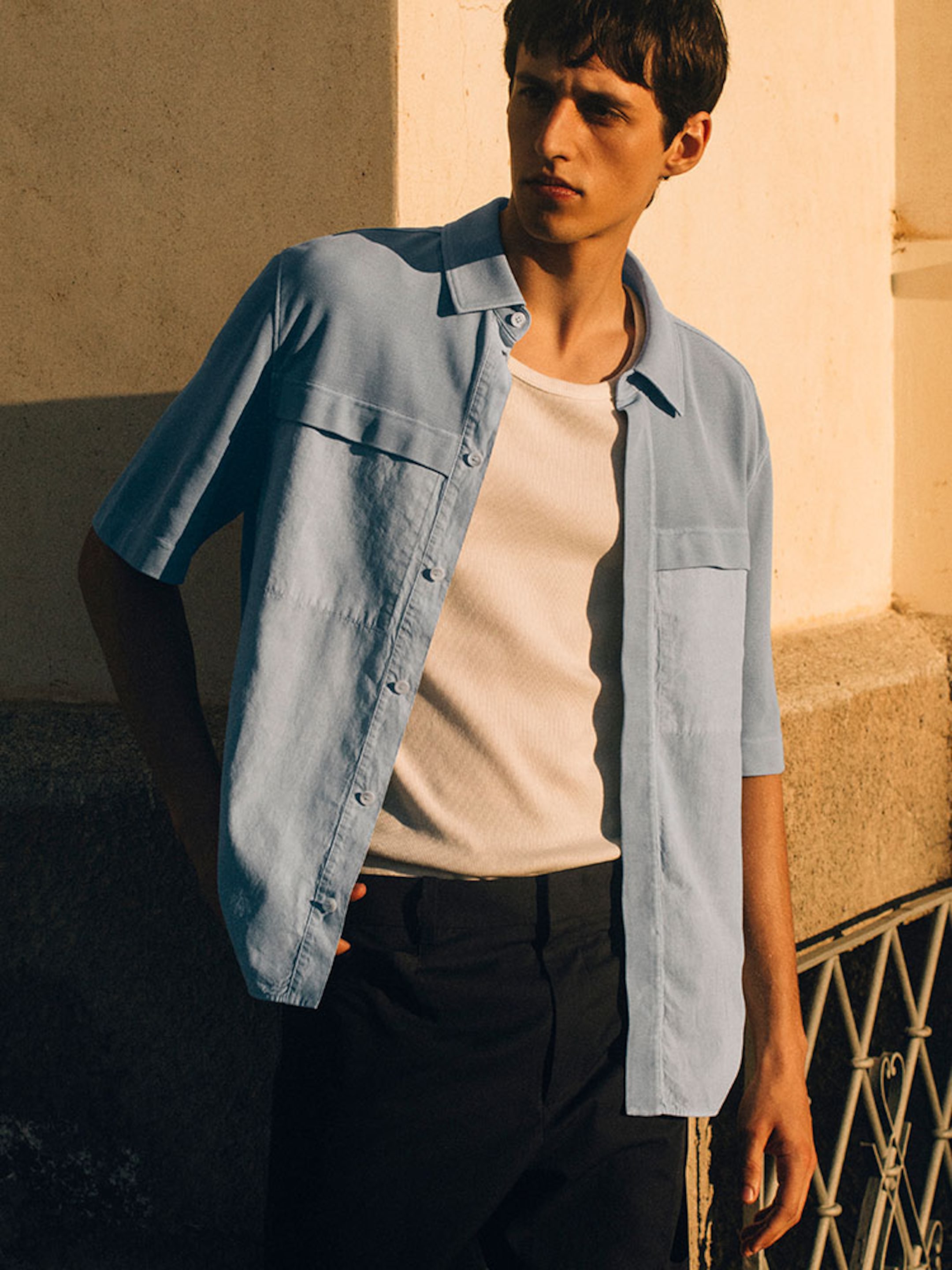 A medium shot of a man standing outdoors in warm, late-afternoon sunlight. He is wearing a light blue, short-sleeved button-down shirt with structured chest pockets over a cream-colored ribbed tank top. The look is paired with black tailored trousers and a slim belt, set against a backdrop of a textured stone building and a decorative metal railing.