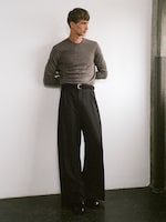 Placeholder for: Person in a gray sweater and wide-leg black pants stands against a white wall with arms behind their back.