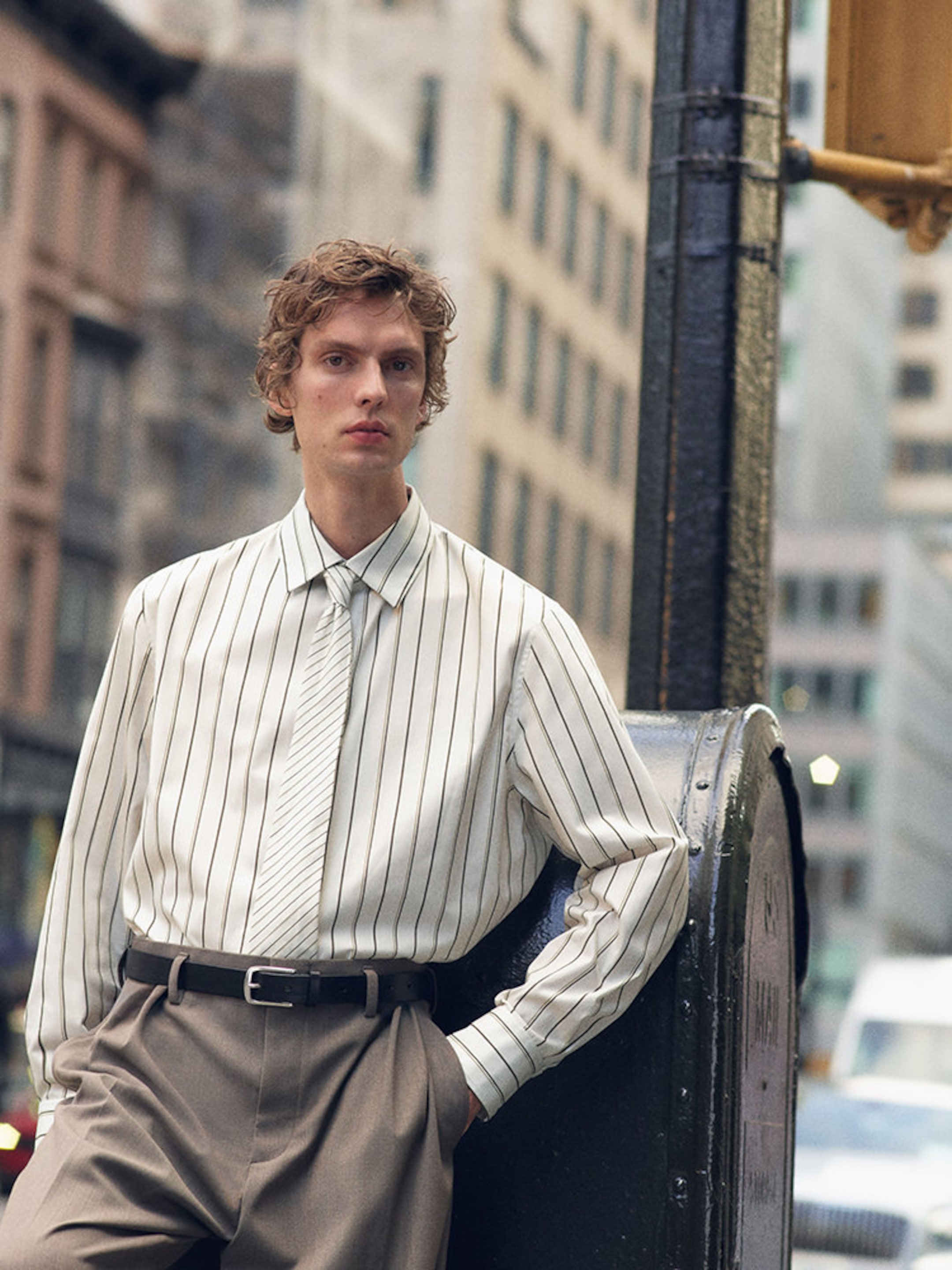 A person in a striped shirt and tie leans against a metal pole on a city street, framed by blurred buildings in the background.