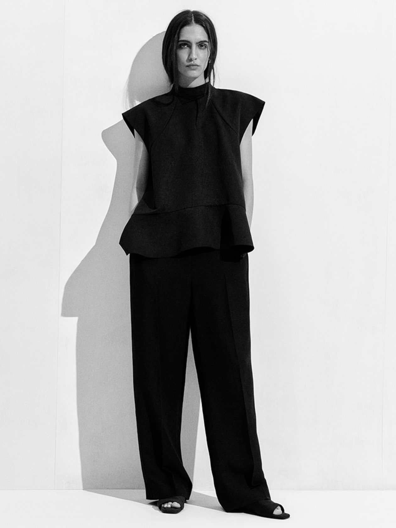 A woman in a sleeveless black outfit stands against a white wall, casting a shadow to her left. Her expression is neutral and her hair is down.