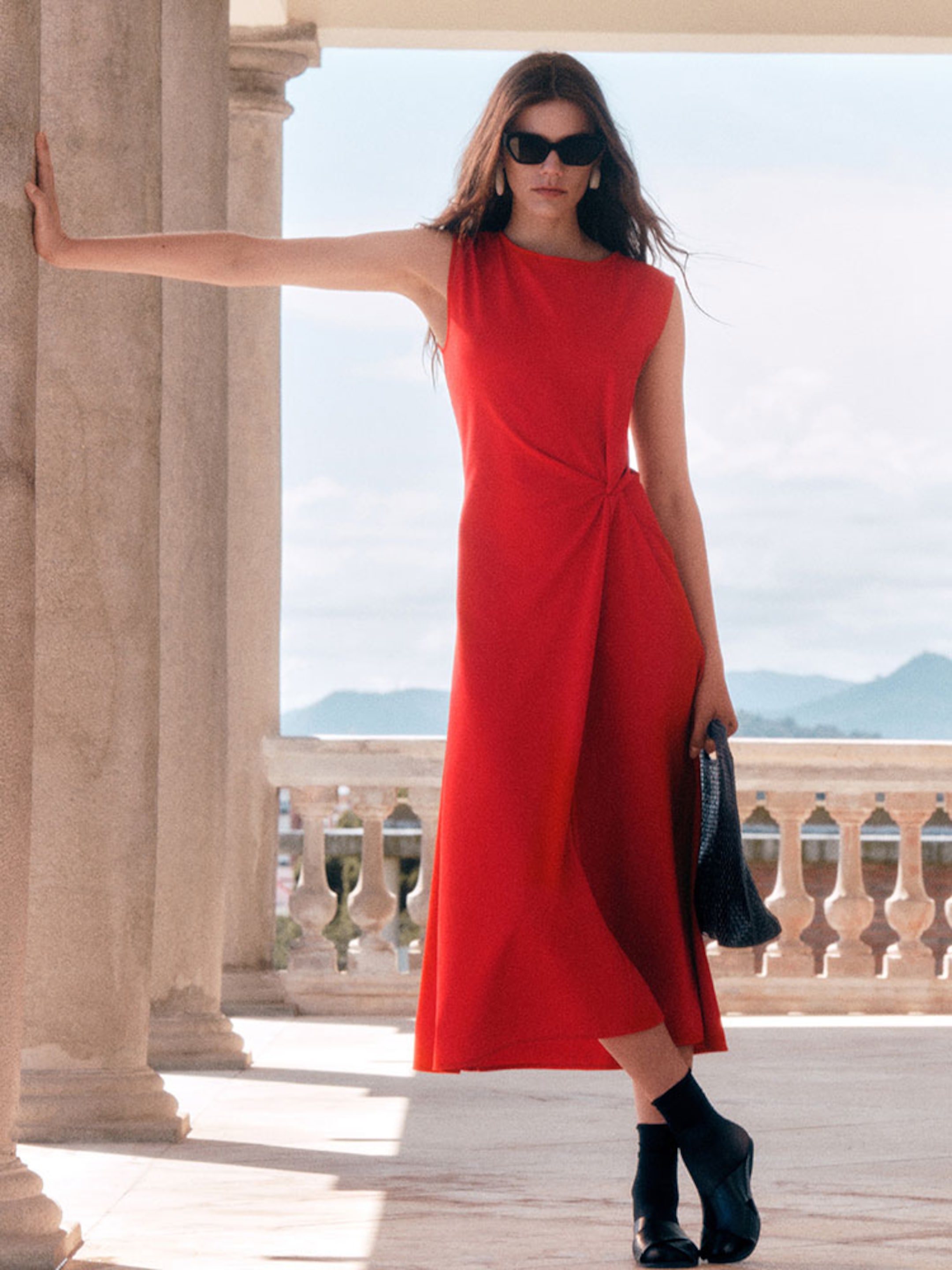 A full-length lifestyle shot of a woman standing and leaning against a stone pillar on a sunlit balcony overlooking a mountain landscape. She is wearing a sleeveless, vibrant red midi dress with a gathered knot detail at the waist, styled with dark sunglasses and black sandals with socks. She carries a dark brown woven leather handbag.