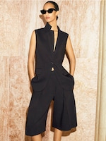 Placeholder for: Person in a sleeveless black suit and wide-leg shorts, wearing dark sunglasses, stands against a marble-textured background.