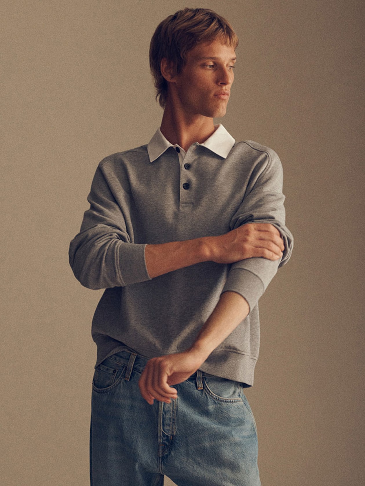 A person wearing a gray pullover with a white collar and blue jeans, looking to the side and adjusting their sleeve, against a neutral background.