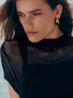 Placeholder for: Person with long hair and gold earrings, wearing a black netted top against a blurred outdoor background.