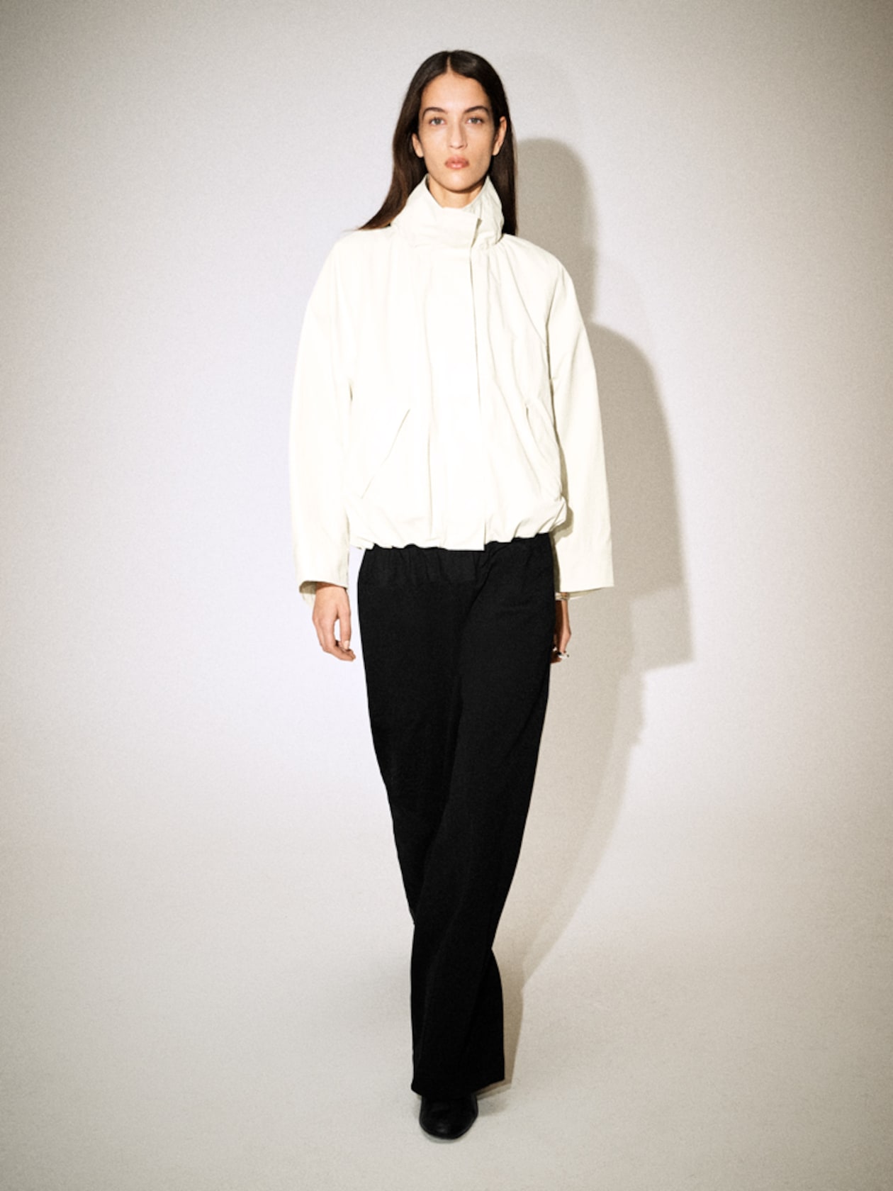 A person wearing a white jacket and black pants walks confidently, casting a shadow on a light background.
