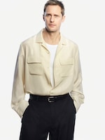 Placeholder for: Man in a cream button-up shirt over a white T-shirt, wearing black pants and belt, stands against a plain white background with hands in pockets.