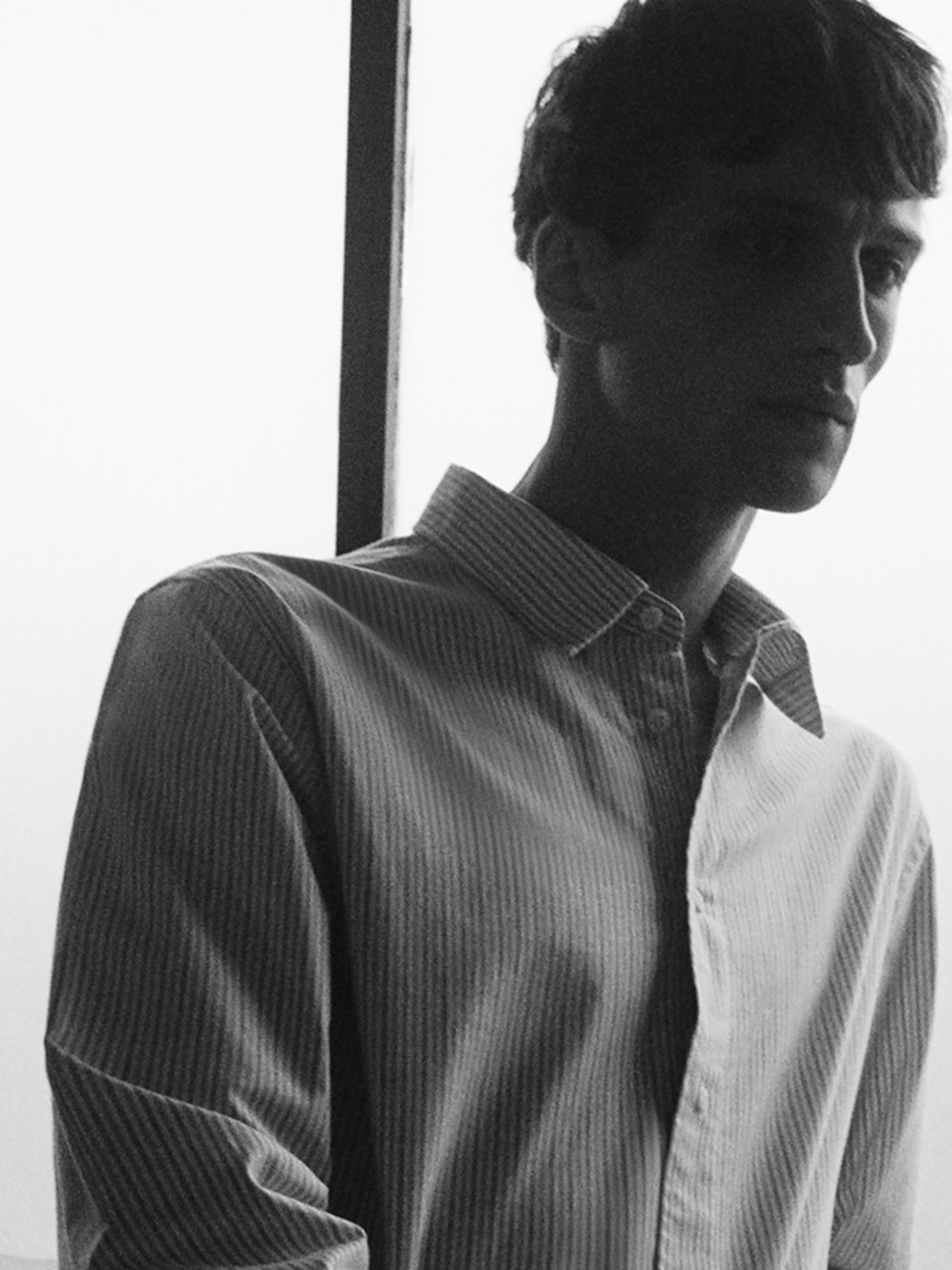 Black and white photo of a person in a striped shirt, standing near a window, looking contemplative with soft lighting.