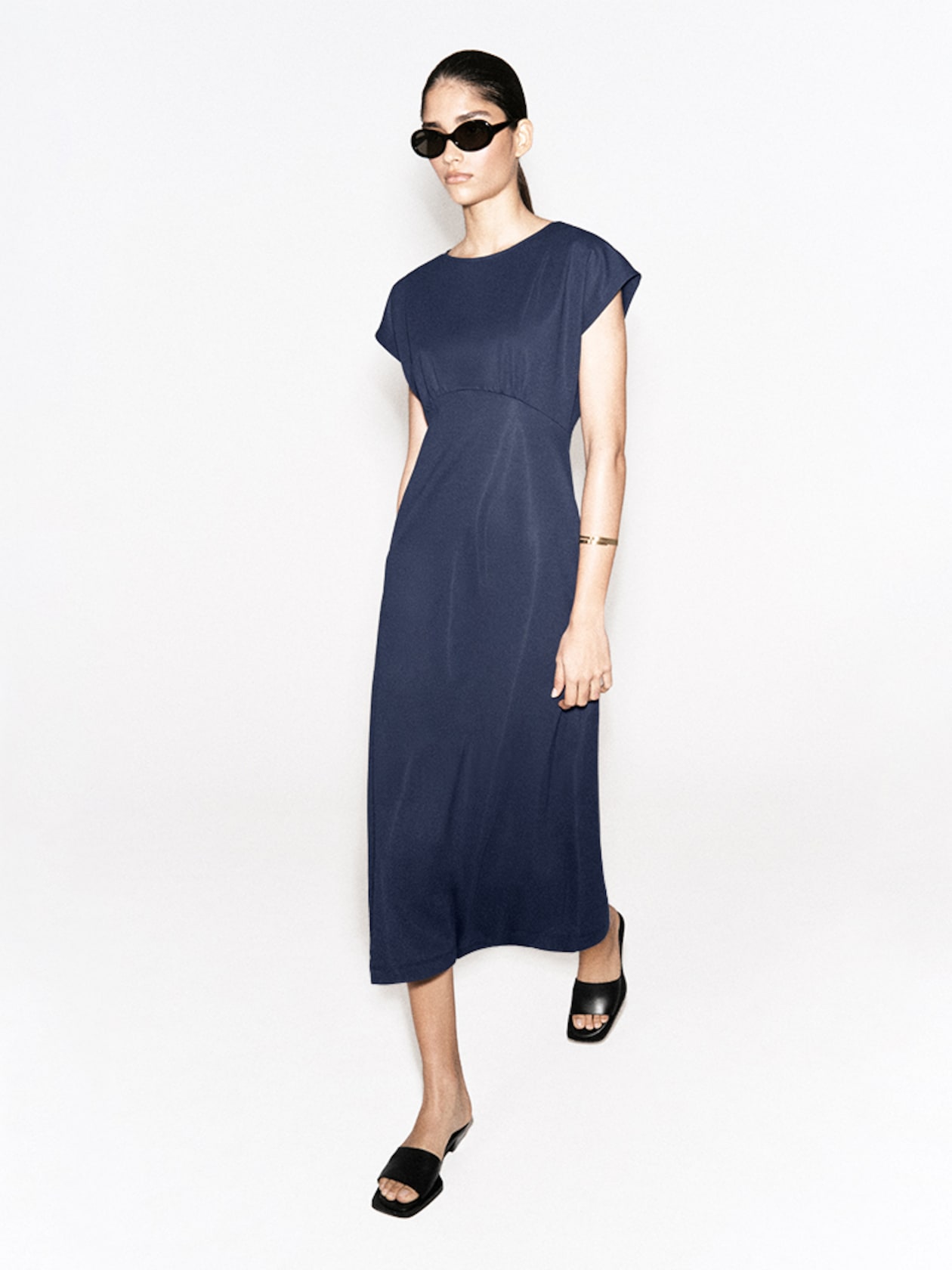 A woman in a navy blue dress and black sandals, wearing sunglasses and a bracelet, stands against a white background.