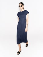 Placeholder for: A woman in a navy blue dress and black sandals, wearing sunglasses and a bracelet, stands against a white background.