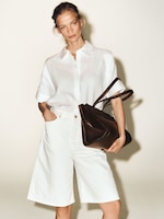 Placeholder for: Woman in a white shirt and shorts, holding a large brown handbag, posing against a plain background.