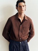 Placeholder for: A man stands against a light background wearing a brown button-up shirt and blue trousers, looking directly at the camera with hands in pockets.