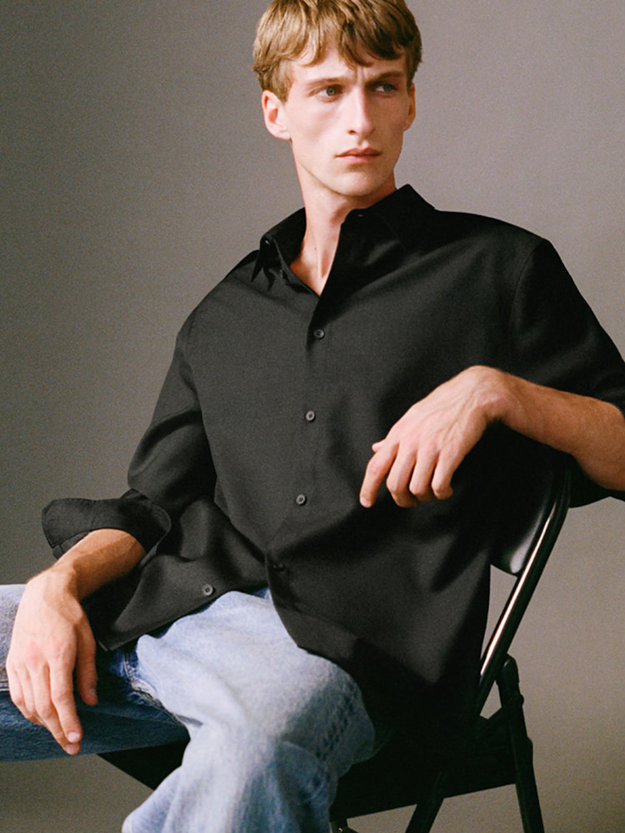 Person sitting on a chair, wearing a black button-up shirt and light blue jeans, looking to the side against a neutral background.
