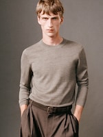 Placeholder for: Person wearing a gray sweater and brown trousers, standing with hands in pockets against a neutral background.