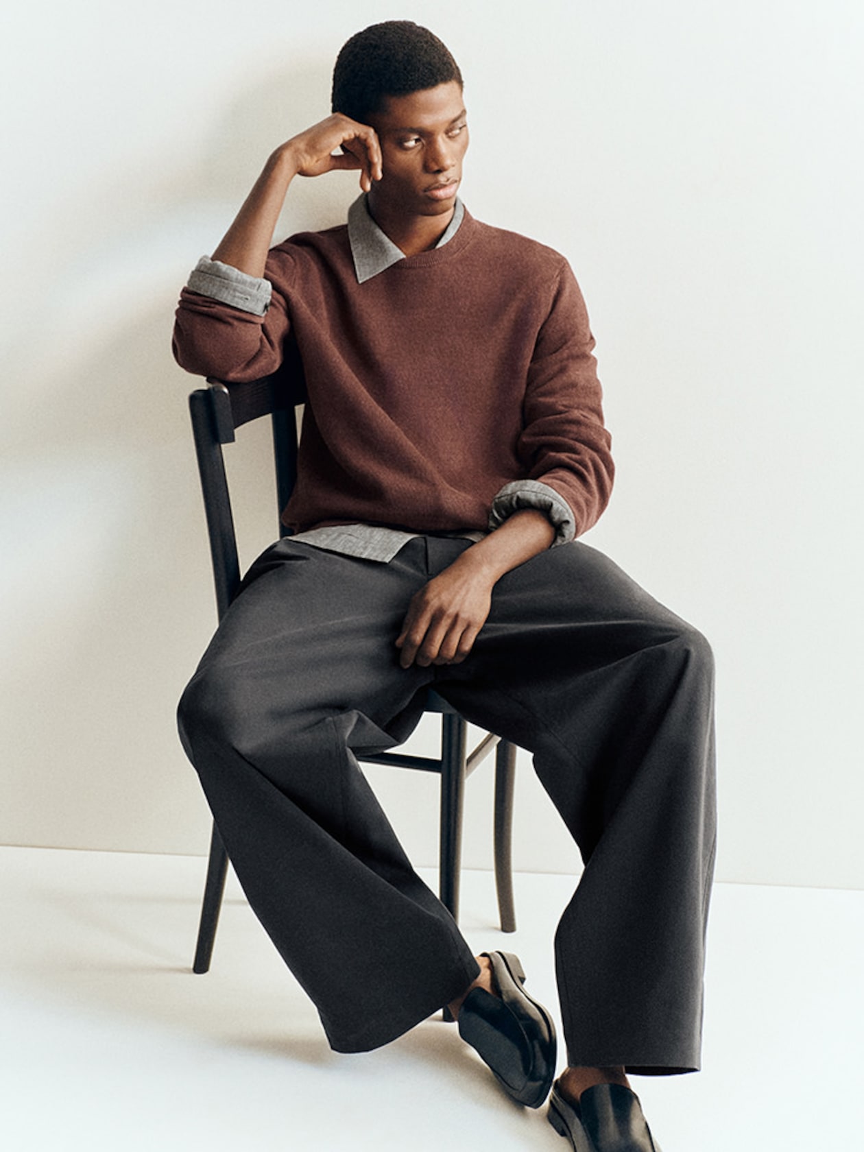 A person sits thoughtfully on a chair wearing a brown sweater, gray shirt, wide gray pants, and black shoes against a plain background.