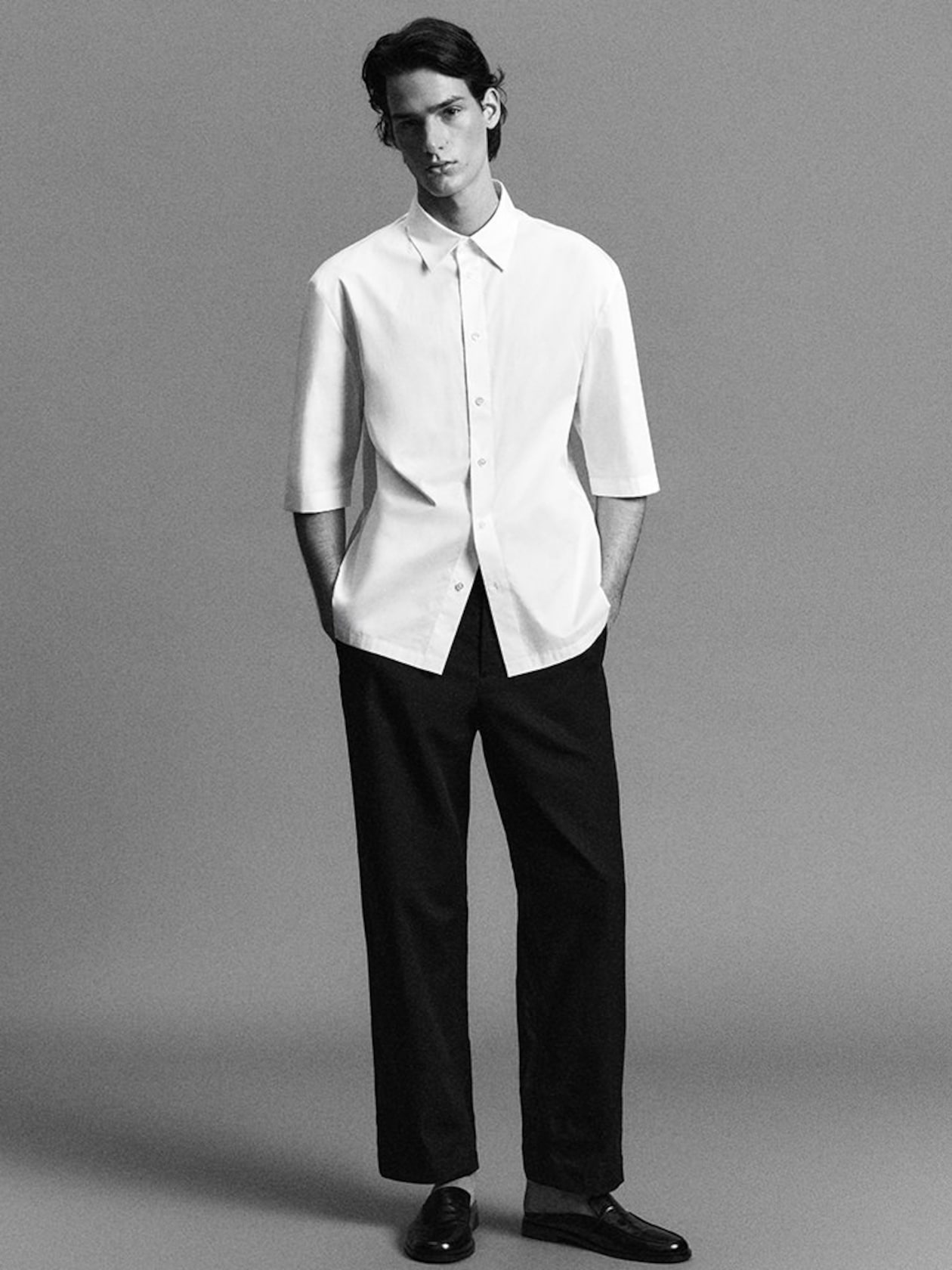 A person wearing a white half-sleeve shirt and black pants, standing against a plain background, hands in pockets, with a neutral expression.