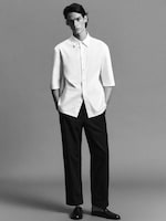 Placeholder for: A person wearing a white half-sleeve shirt and black pants, standing against a plain background, hands in pockets, with a neutral expression.