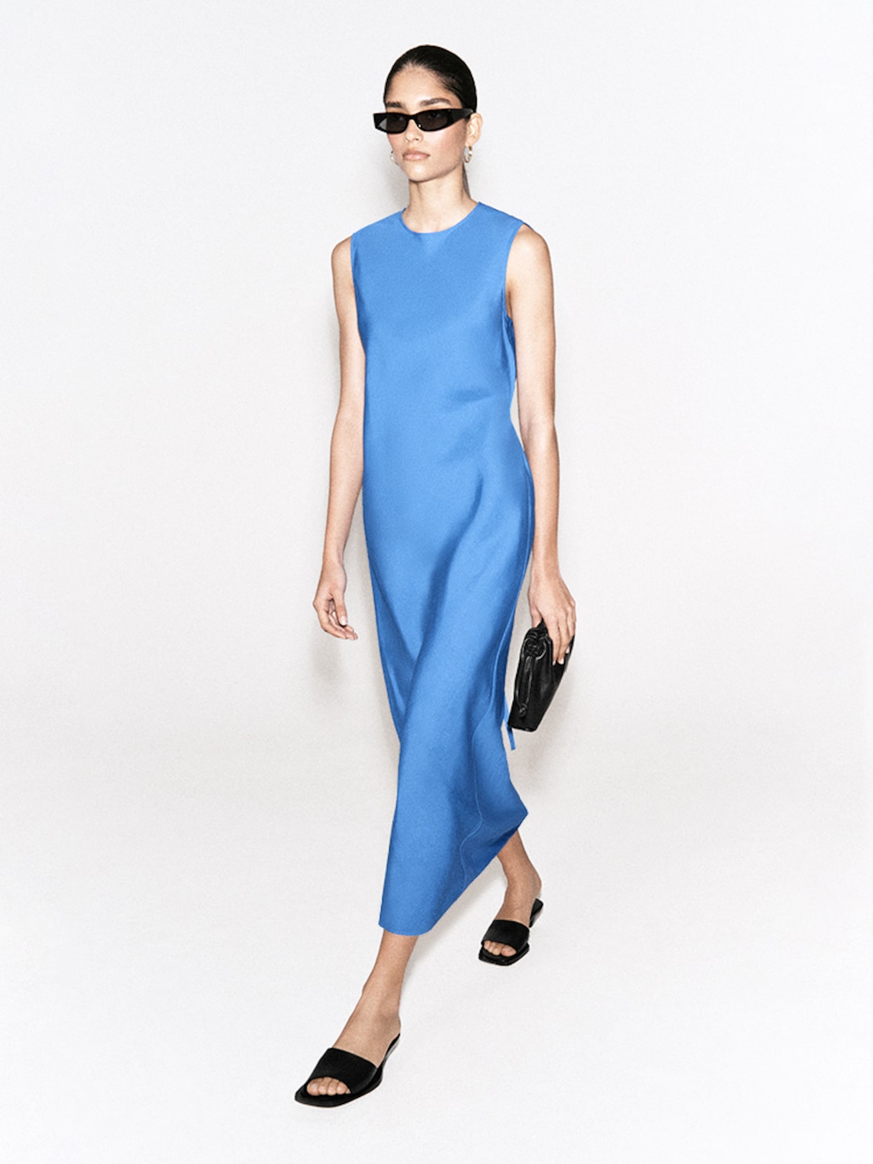 A person wearing sunglasses and a sleeveless blue dress walks while holding a black clutch, sporting black slide sandals.