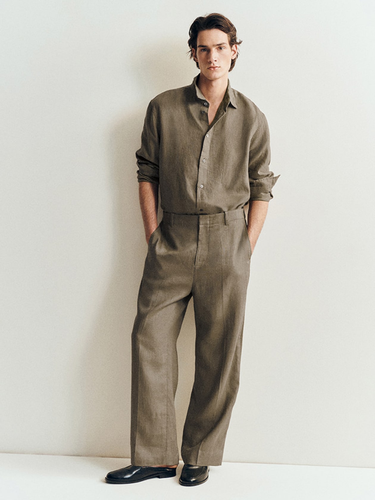 A man wearing a loose-fitting, brown shirt and pants with black shoes stands against a light-colored wall, hands in pockets, looking at the camera.