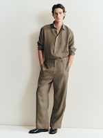 Placeholder for: A man wearing a loose-fitting, brown shirt and pants with black shoes stands against a light-colored wall, hands in pockets, looking at the camera.