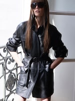 Placeholder for: A woman with long hair wears a belted black leather trench coat and oversized sunglasses, standing by a decorative railing in soft natural light.