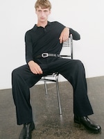 Placeholder for: Person sitting on a metal chair, wearing a black long-sleeve shirt, black pants, and boots, against a white wall and concrete floor.