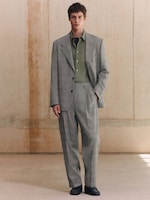 Placeholder for: A person wearing a gray suit with a green shirt stands in a minimalist room with beige walls.