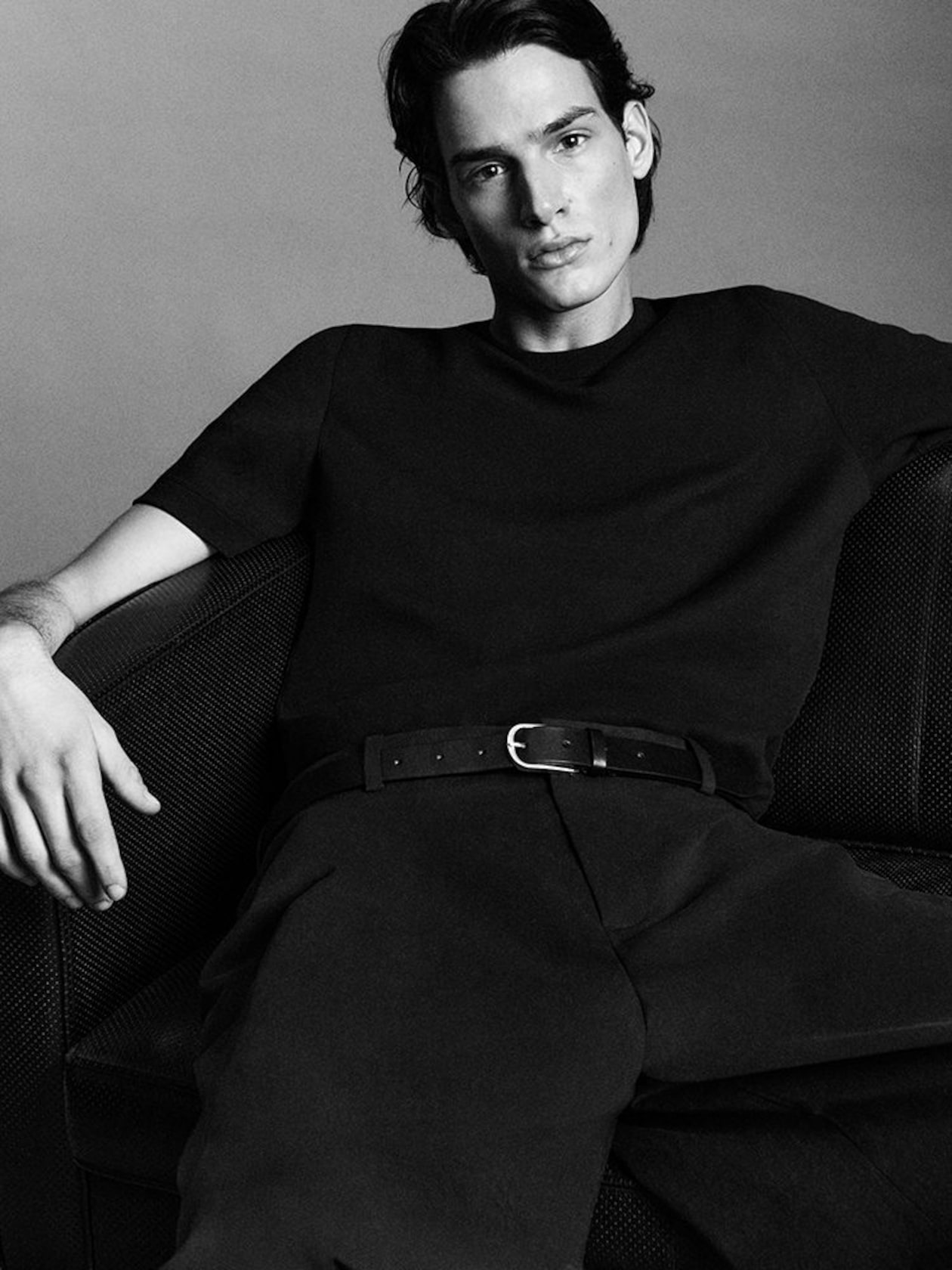 A person in a black outfit sits on a couch, looking directly at the camera with a relaxed posture, in a black-and-white photograph.