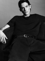 Placeholder for: A person in a black outfit sits on a couch, looking directly at the camera with a relaxed posture, in a black-and-white photograph.