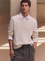 Placeholder for: Person wearing a cream textured sweater over a white shirt and gray trousers, standing against a pillar in soft, natural lighting.