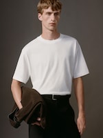 Placeholder for: Person in a white t-shirt holding a dark jacket over their arm, standing against a plain dark background.