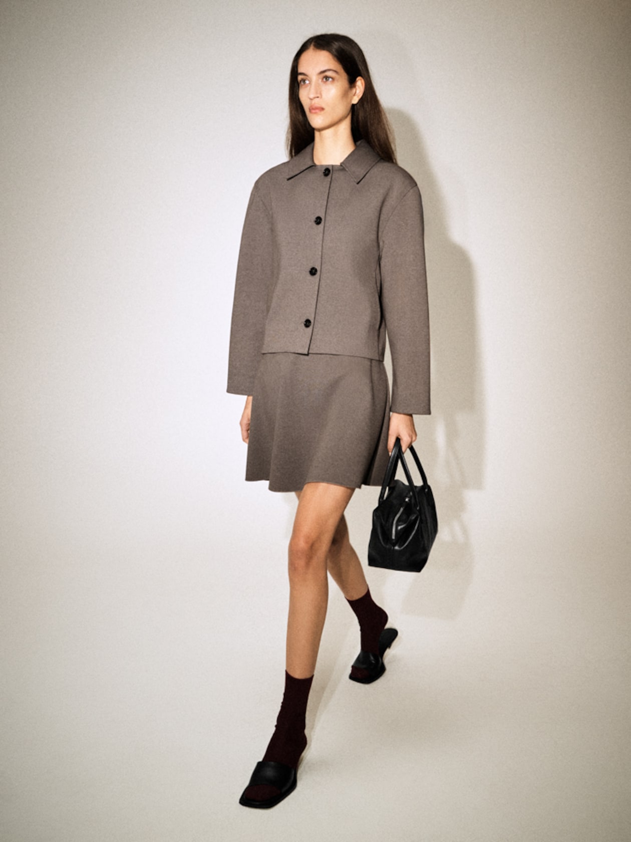 A woman in a gray jacket and skirt, wearing maroon socks and black sandals, walks while holding a black handbag against a plain background.
