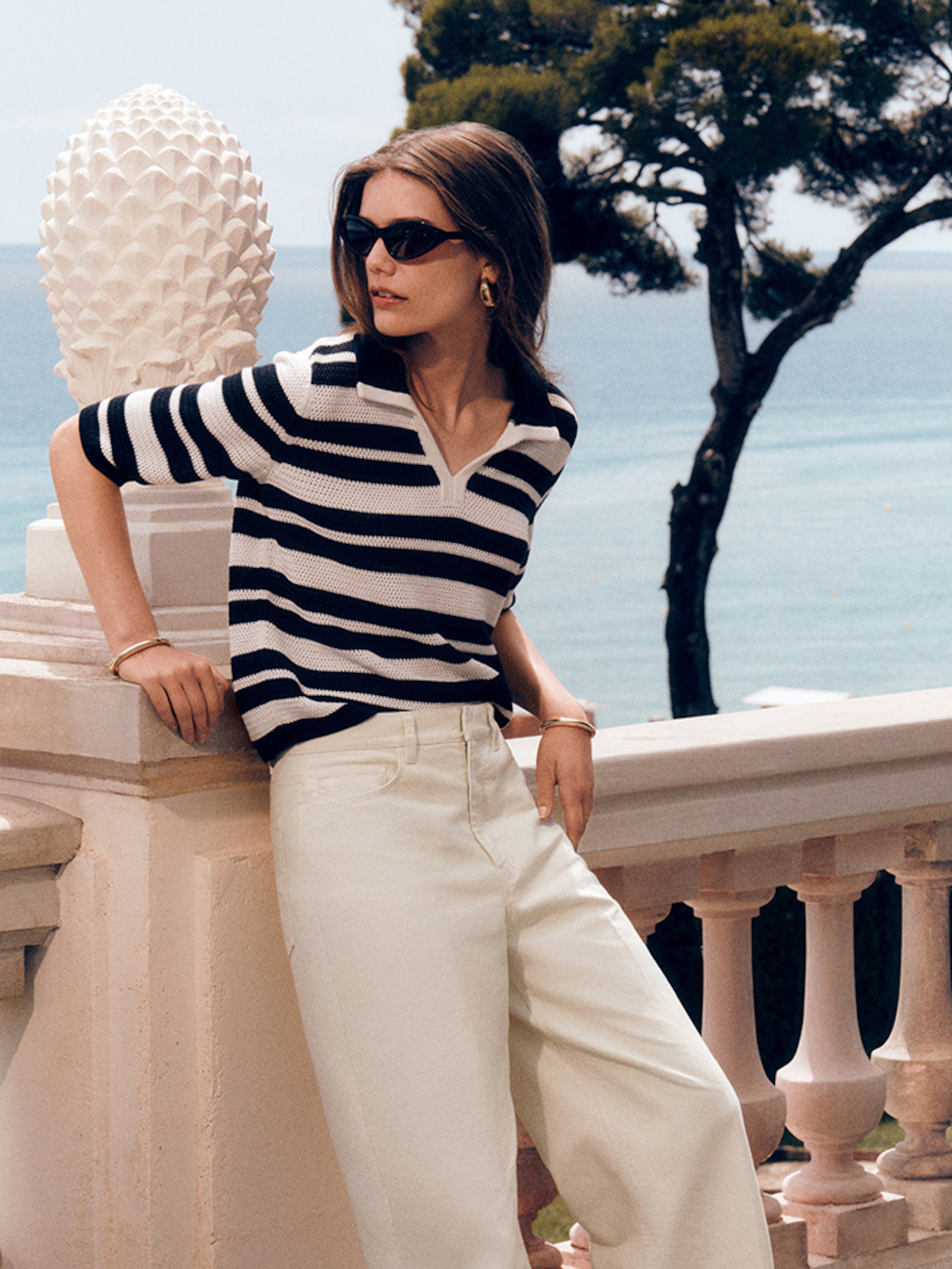 A woman in sunglasses leans on a balustrade, wearing a striped top and white pants, with the ocean and a tree in the background.