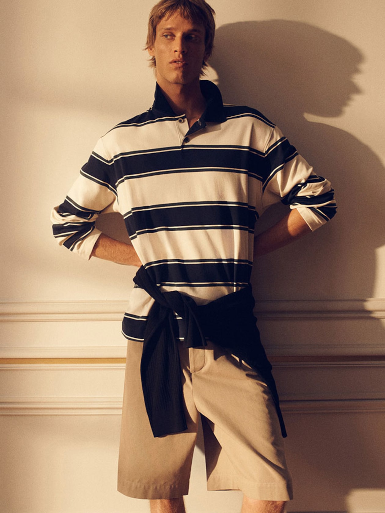 Man wearing a striped polo and khaki shorts, with a sweater tied around his waist, standing indoors against a wall with soft lighting.