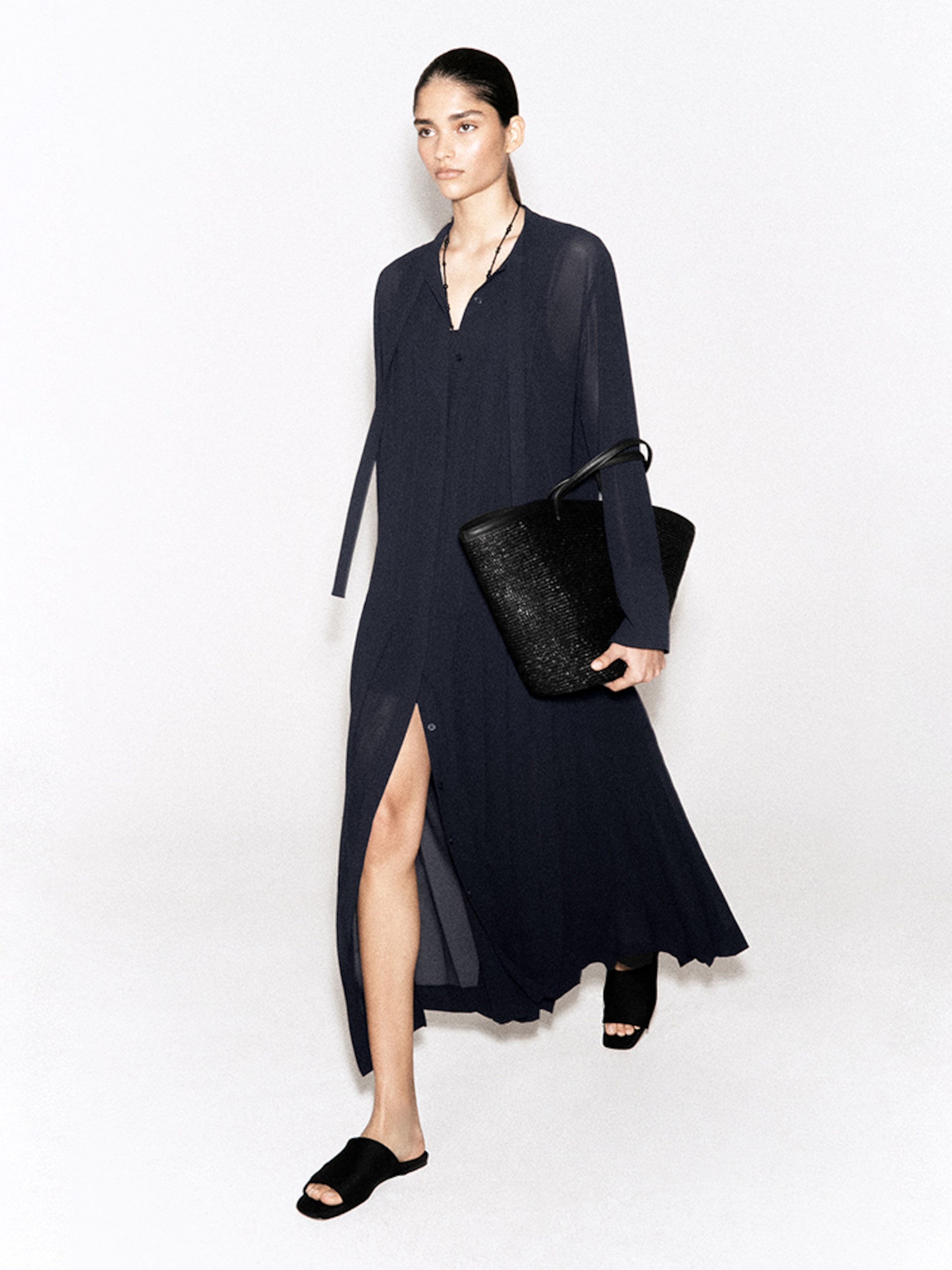 A person wearing a long, dark blue dress with a slit, black sandals, and holding a large black textured tote bag, walking against a white background.
