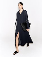 Placeholder for: A person wearing a long, dark blue dress with a slit, black sandals, and holding a large black textured tote bag, walking against a white background.