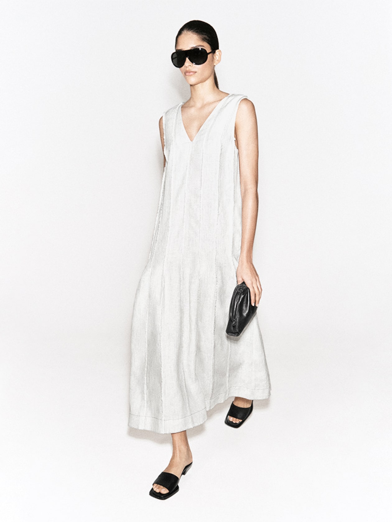 A woman in a sleeveless, long white dress wears large black sunglasses, carrying a small black clutch, and walking in black heeled sandals.