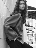Placeholder for: Black and white photo of a person with long hair leaning against a wall on a city street, wearing a loose, long-sleeved top and pants.
