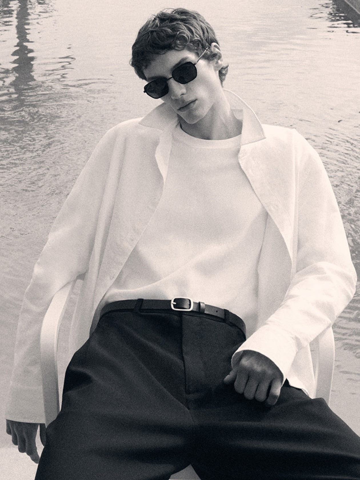 Person in sunglasses and white shirt lounging by water, wearing dark pants with a belt, exuding a relaxed and stylish vibe.