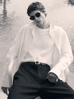 Placeholder for: Person in sunglasses and white shirt lounging by water, wearing dark pants with a belt, exuding a relaxed and stylish vibe.