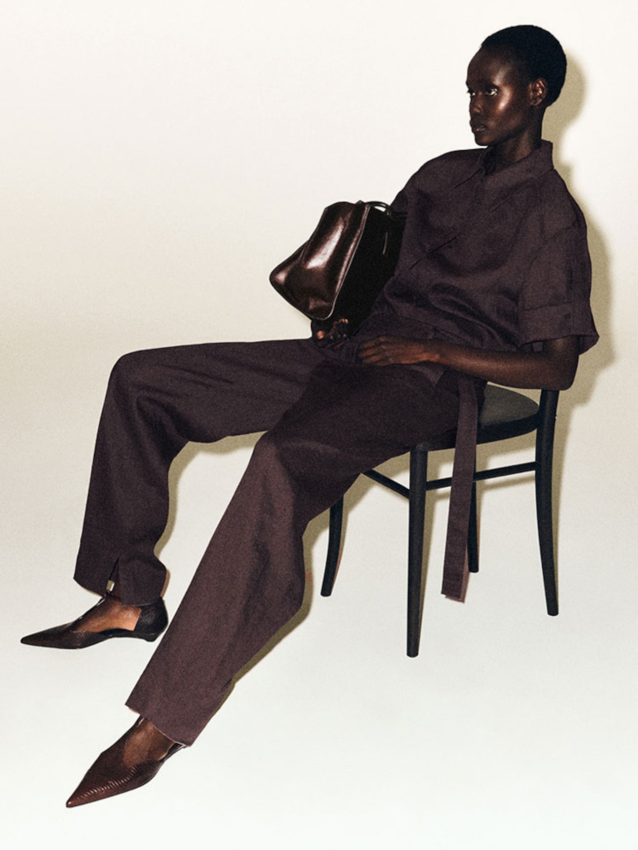 A person in a dark brown outfit sits on a chair, holding a brown bag, with legs casually stretched out and wearing pointed shoes.